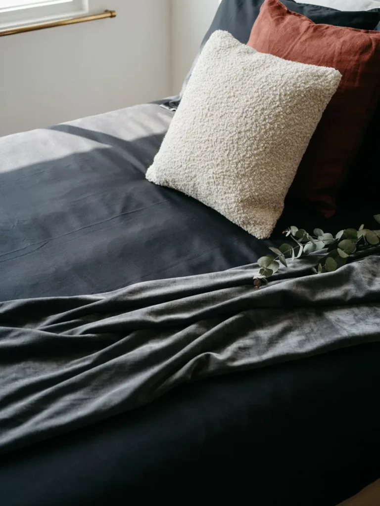 A close up of layered bedding showing black satin sheets, a velvet throw, and mixed texture pillows