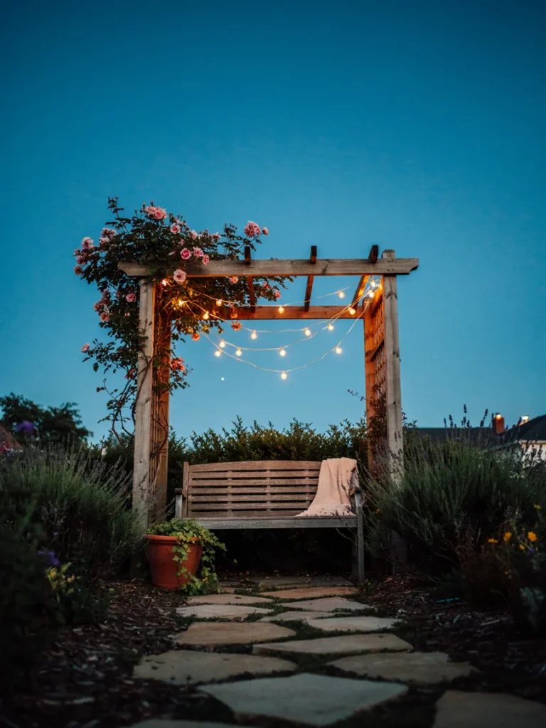 A garden arbor strung with warm white string lights at dusk, with a wooden bench underneath]