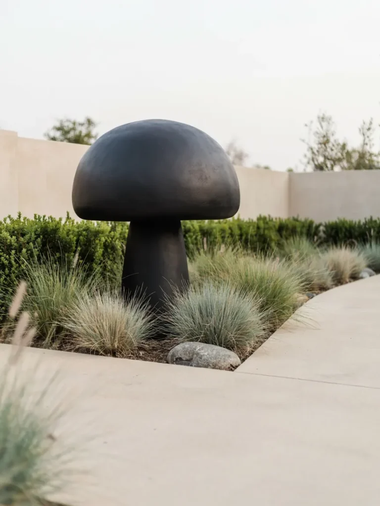 A simple white or matte black mushroom sculpture set against clean hardscaping and ornamental grasses in a contemporary garden