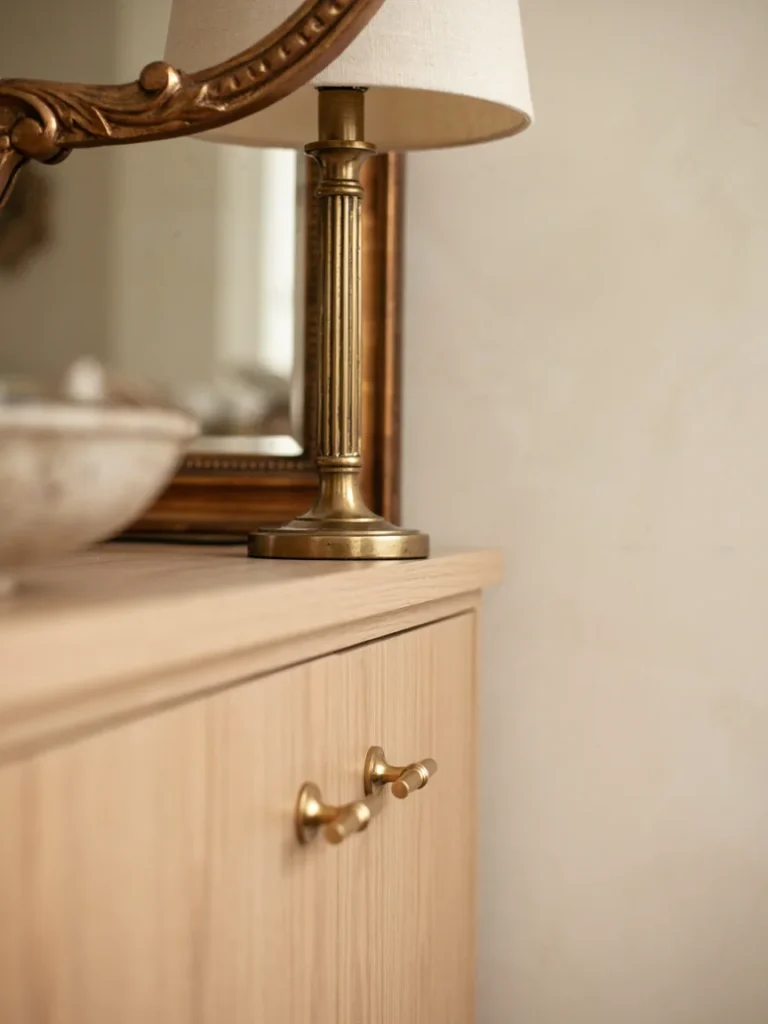 Close up of brass and gold metallic accents, a mirror frame, lamp fixture, and drawer pulls, warm elegant metallic tones against a soft neutral background