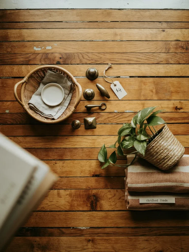 Flat lay of affordable secondhand decor finds, a small rattan basket, vintage brass drawer pulls, a folded earth tone throw, and a leafy plant in a woven planter, budget styling on a wooden surface