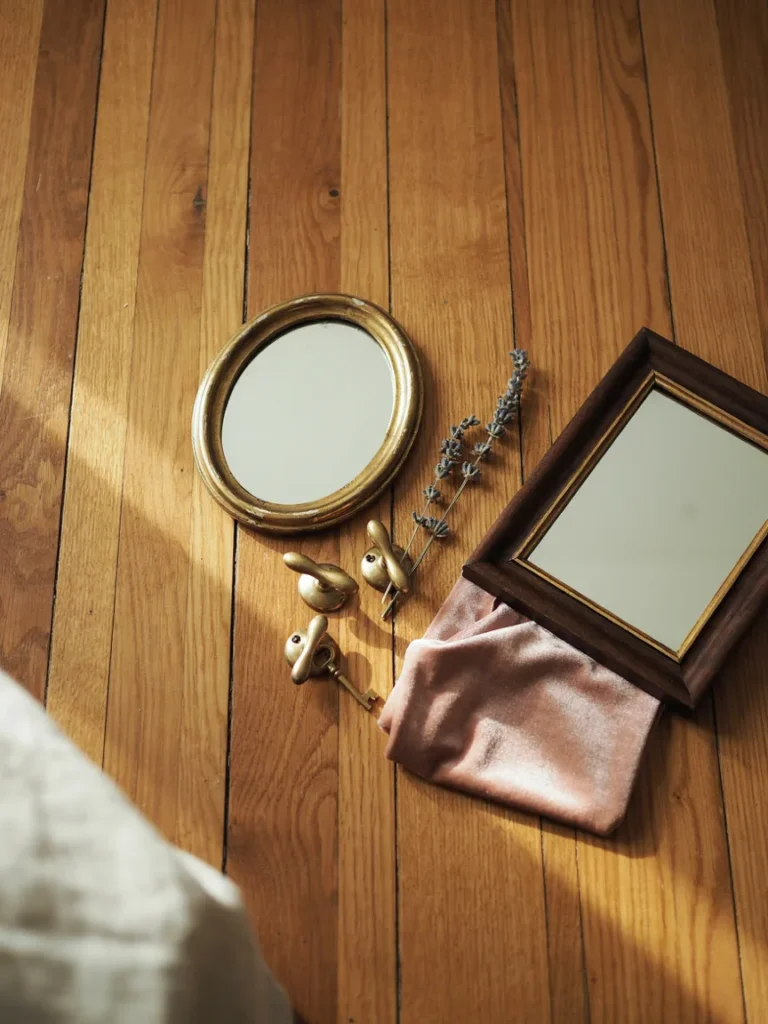 Flat lay of budget friendly Parisian decor finds, a small ornate secondhand mirror, brass drawer pulls, a folded blush velvet fabric swatch, and a vintage picture frame, styled on a wooden surface