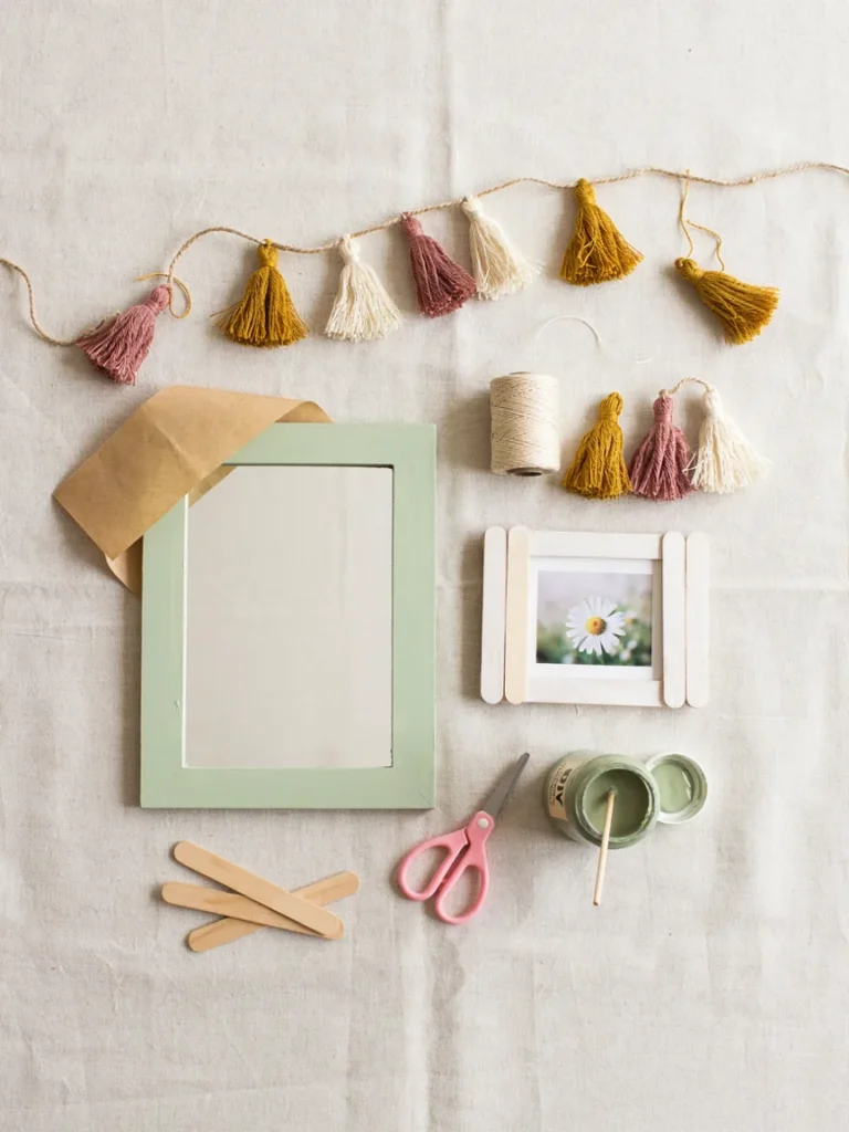 Flat lay of budget friendly beginner DIY pieces, a painted mirror frame, yarn tassel garland, and popsicle stick picture frame, simple approachable craft styling