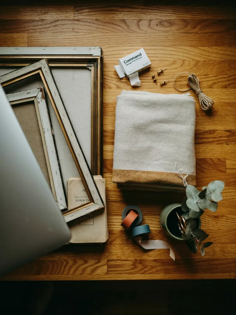 Flat lay of budget friendly college decor finds, secondhand frames, a folded thrifted throw blanket, and command strips, styled on a wooden surface, affordable dorm and apartment decor materials
