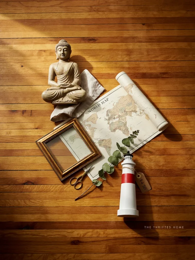 Flat lay of budget friendly themed decor finds, a small secondhand Buddha figure, a printed map ready for framing, and a simple model lighthouse, styled on a wooden surface