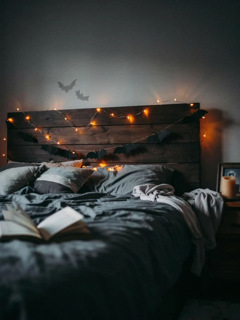 Orange tinted string lights draped along a headboard with a black paper bat garland strung nearby, warm eerie glow, spooky bedroom lighting setup