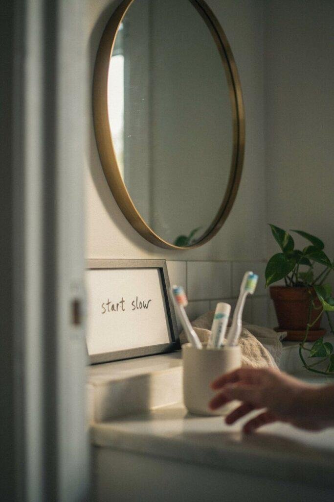 Close up of a small framed quote print positioned beside a bathroom mirror, warm morning light, person's blurred hand reaching for a toothbrush in the foreground, cozy everyday bathroom moment