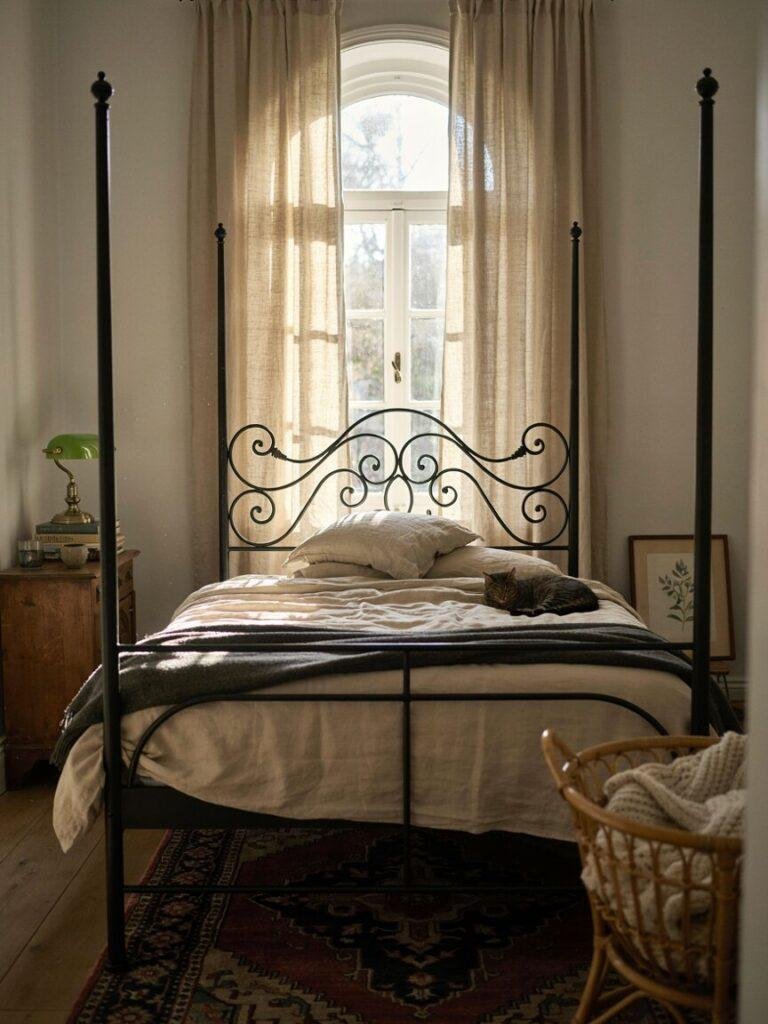 A styled photo of a black wrought iron or four poster bed frame as the room's centerpiece, shot from the foot of the bed